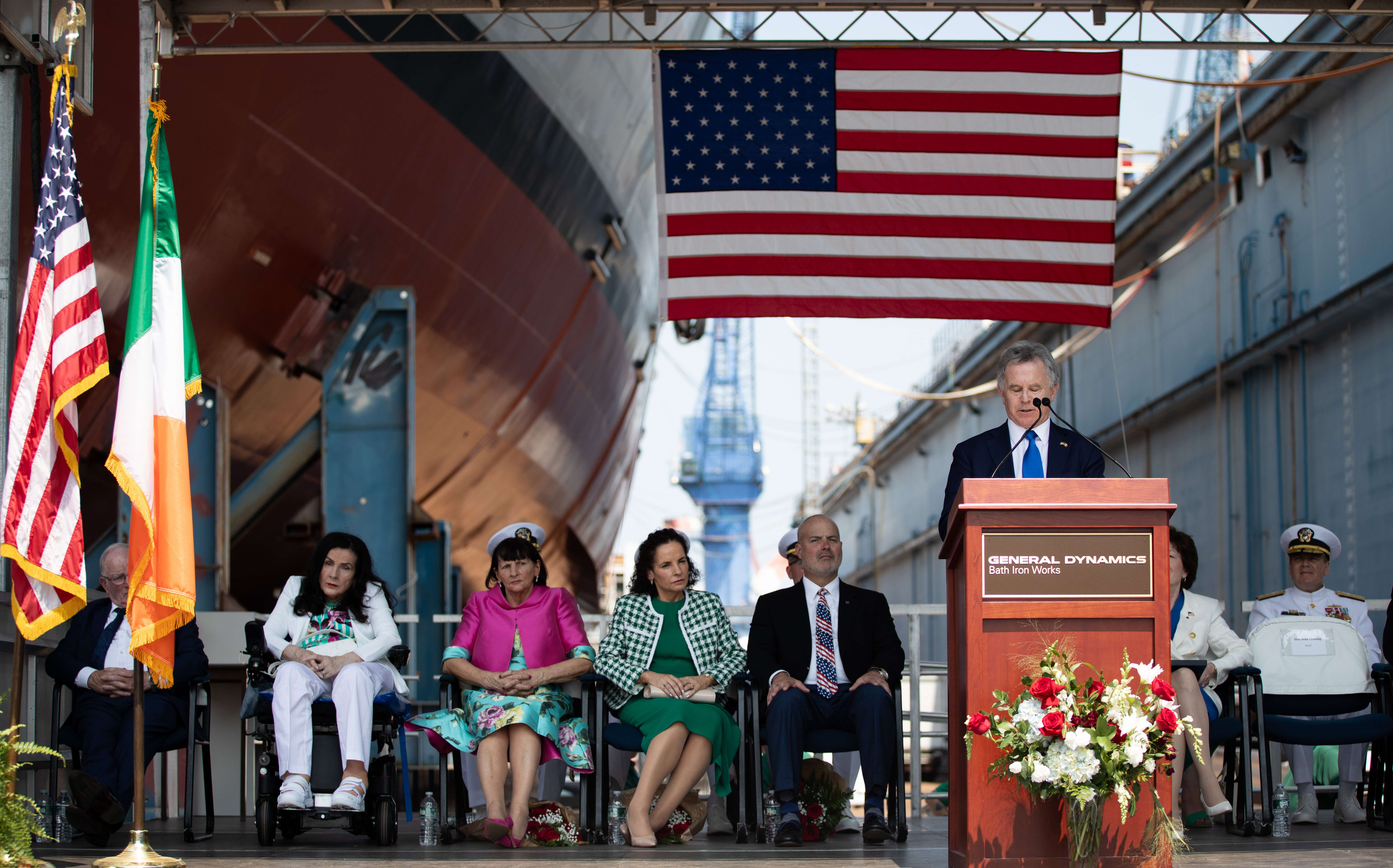 People seated and one standing at a podium at BIW' ship christening.