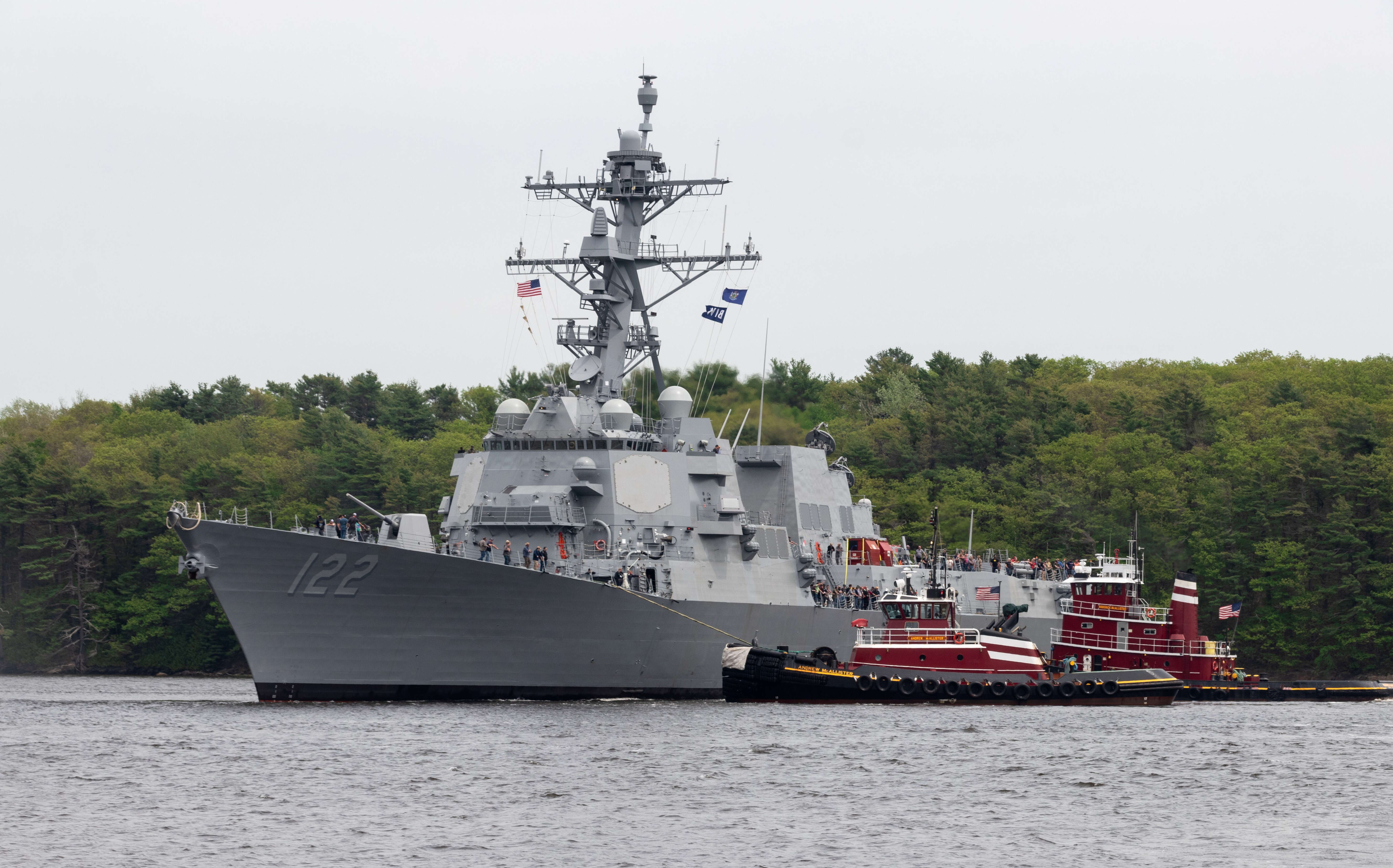 A gray warship and tugboats are on the water.