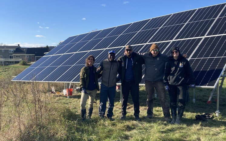 Five people stand in a field in front of solar panels.