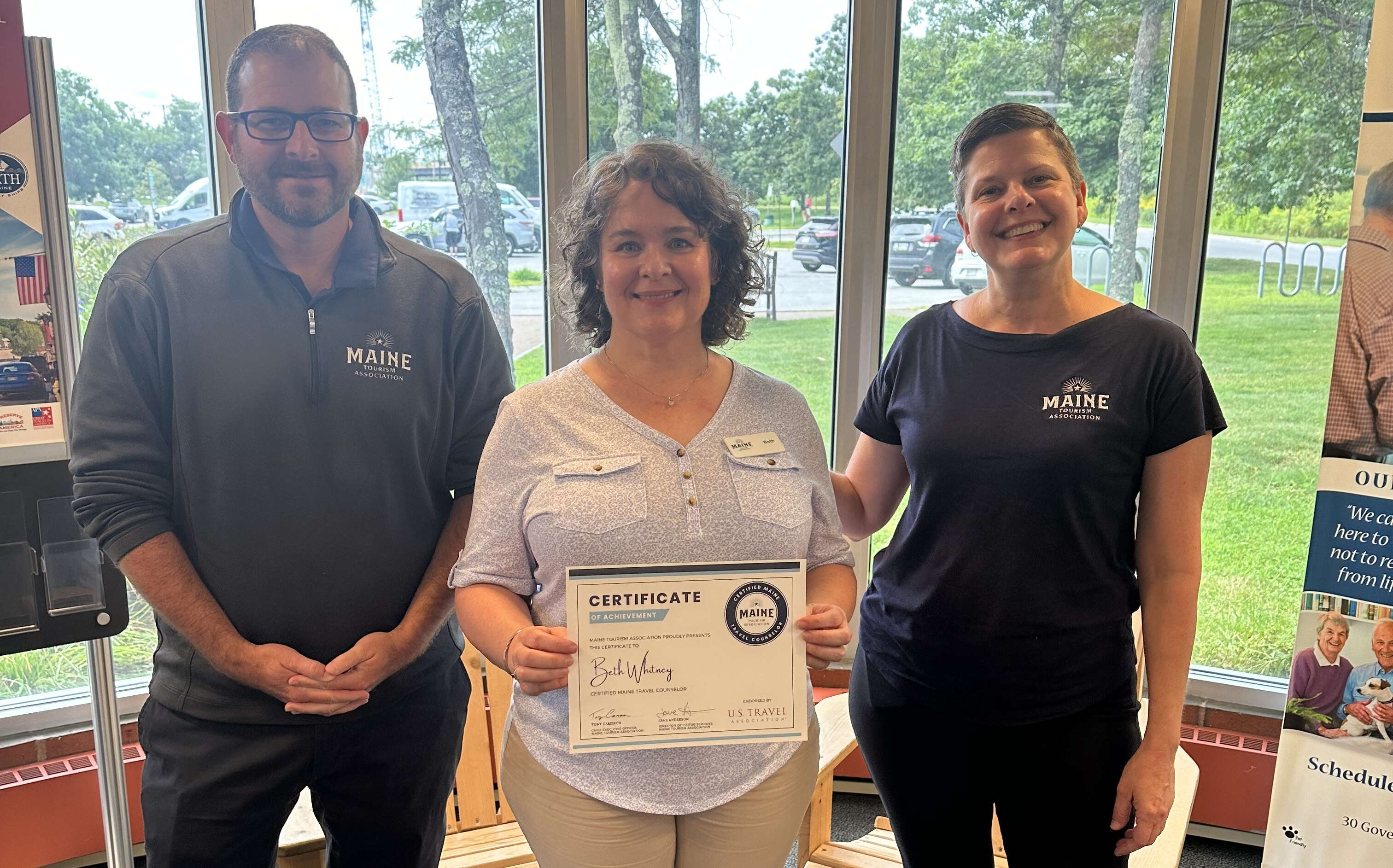 Three people pose with a certificate in front of big windows.