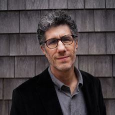 portrait of man with glasses and black jacket, in front of gray clapboards