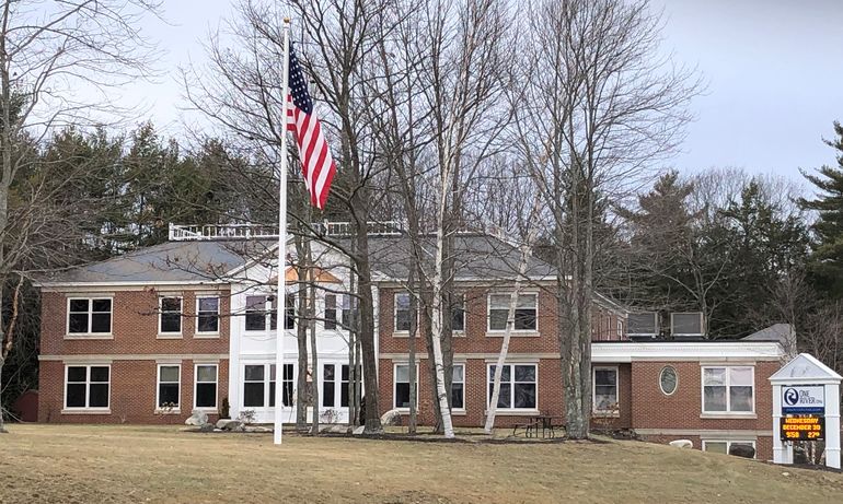 a two-strory large brick building with white trim, with a green lawn and woods in the background and an american flag on a pole in front