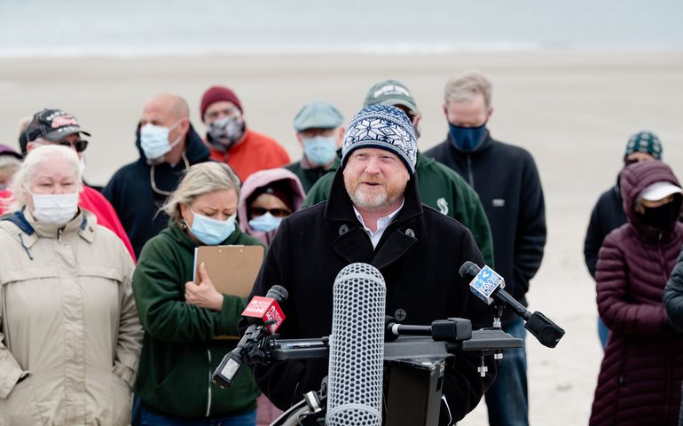 a man, white with a beard, wearing a ski cap, speaks into a microphone on a cold windy beach and several people in coats and wearing face coverings stand behind him