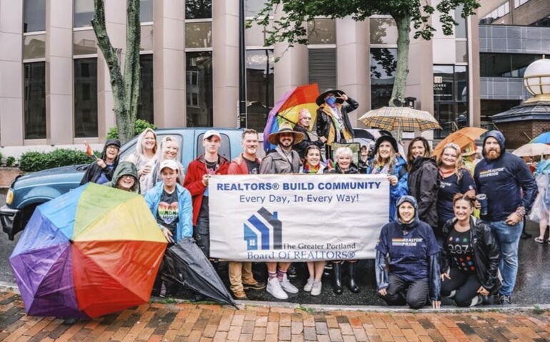 A group of people pose with a banner and colorful umbrellas outside on a brick sidewalk apparently in the rain.