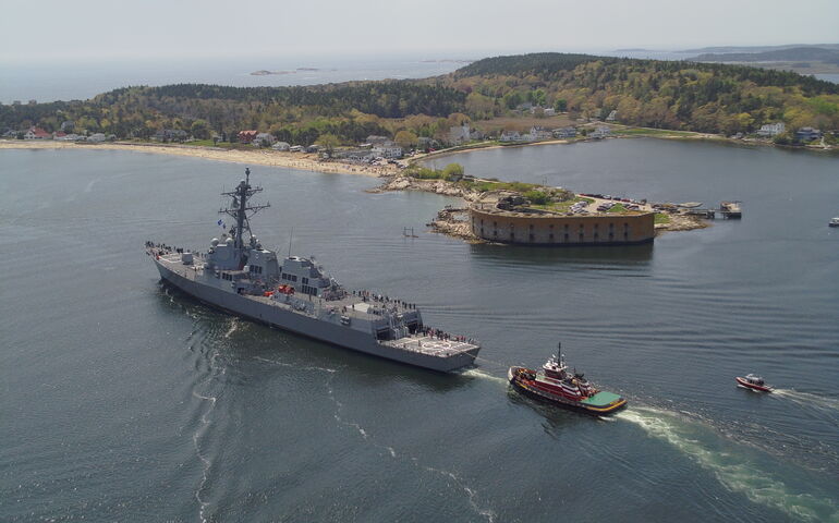 A large gray ship is seen from the air cruising through a bay with a couple of smaller ships behind it.