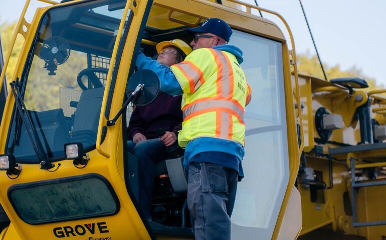 A person sits in a crane cabin while another person looks on.